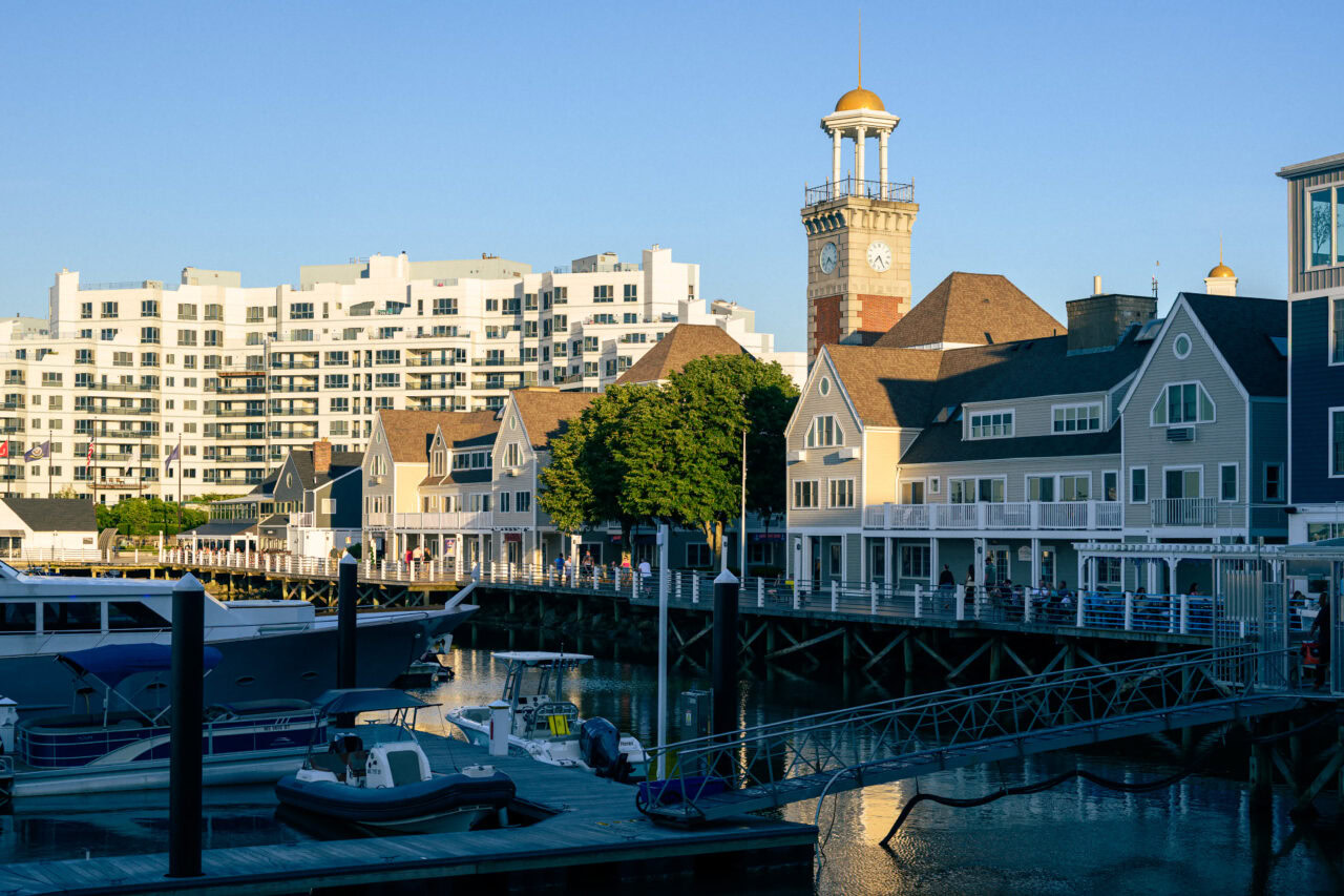 Marina Bay waterfront in Quincy, Massachusetts at golden hour, showing boats in the marina, waterfront buildings, and people on the boardwalk.