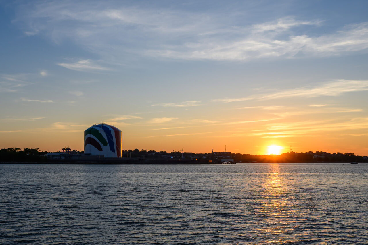 Painted LNG storage tank silhouetted against a vivid orange and blue sunset sky over the harbor from Squantum Point Park in Quincy, Massachusetts, with the setting sun reflected on the water.