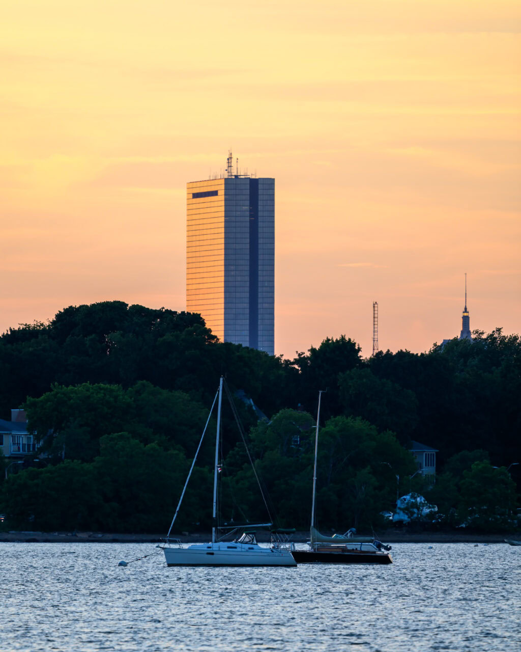 Two sailboats moored on the harbor at Squantum Point Park in Quincy, Massachusetts, with the John Hancock Tower catching the last orange light of sunset above a dark tree line.