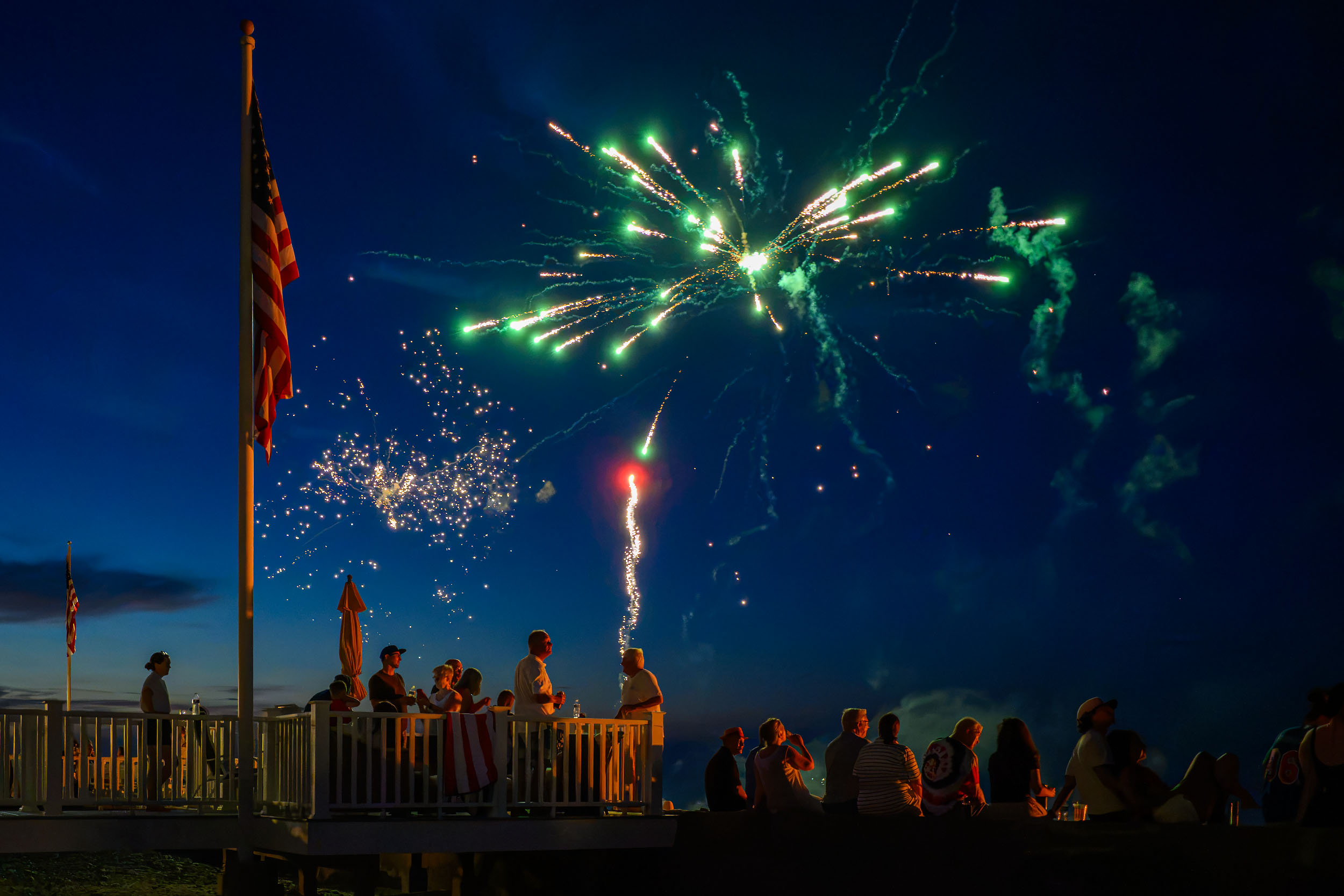 People gathering on a deck under green fireworks during blue hour in Humarock, Scituate.